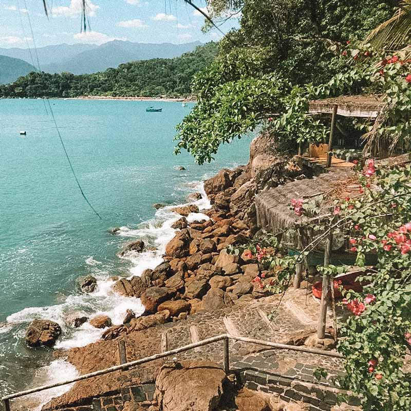 Casa bem na costa litorânea, com algumas pedras e um passarela com corredor dando acesso direto ao mar. Ao redor da casa, a vegetação da Mata Atlântica