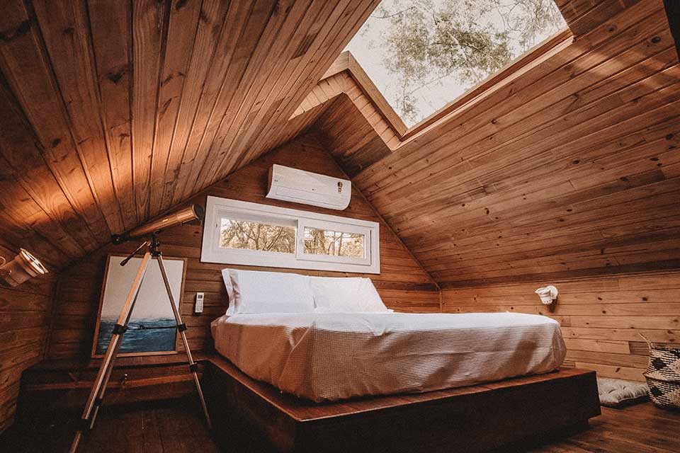 Quarto todo em madeira, com cama de casa e teto de vidro acima da cama. Ao lado da cama, um telescópio.