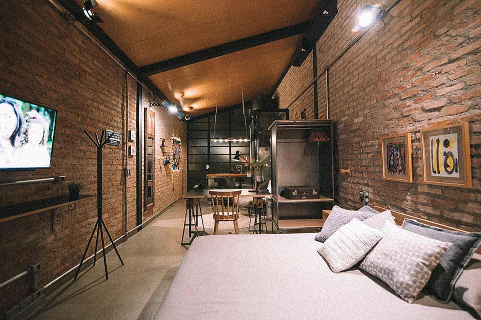 Loft industrial em SP. EM primeiro plano uma cama de lençol branco em frente à TV. Ao fundo, a cozinha e banheiro do apê.