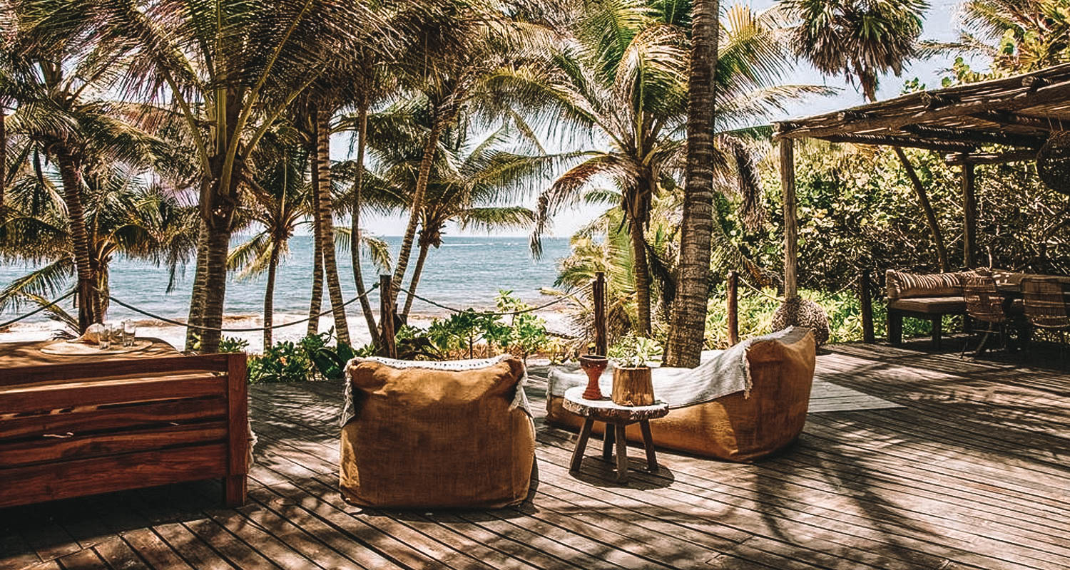 Deck de madeira com poltronas, sala de jantar exposta, palmeiras e o mar a frente
