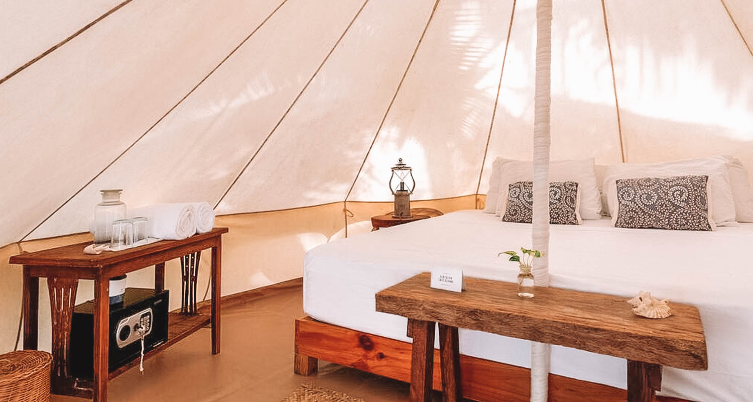 Dentro da barraca, cama de casal ao meio e mesinha de madeira ao lado com toalhas e cofre