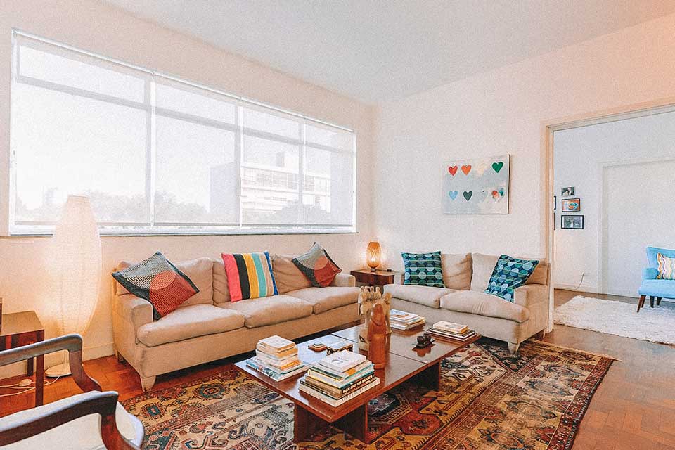 Sala de estar com dois sofás, almofadas coloridos, uma mesa de centro com objetos decorativos e amplas janelas.