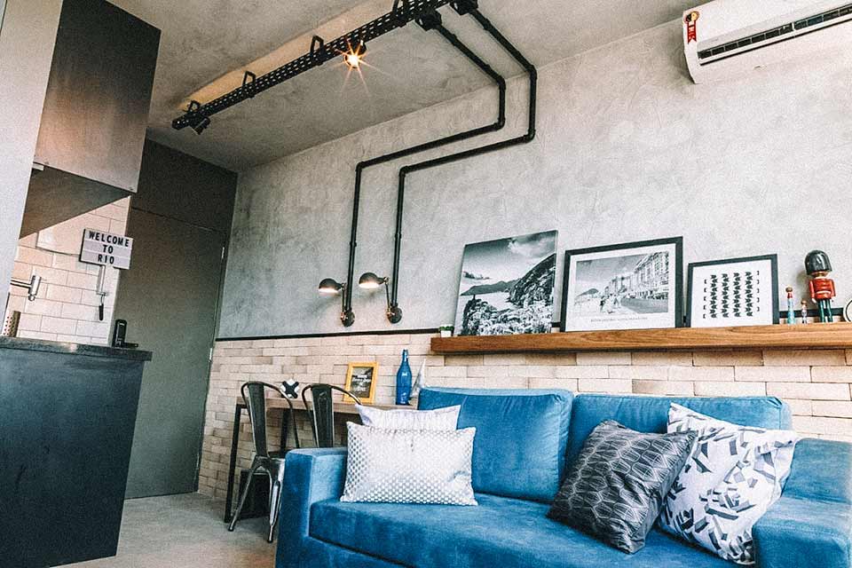 Loft com sofá azul, quadros encostados na prateleira, parede de cimento queimado e ar-condicionado