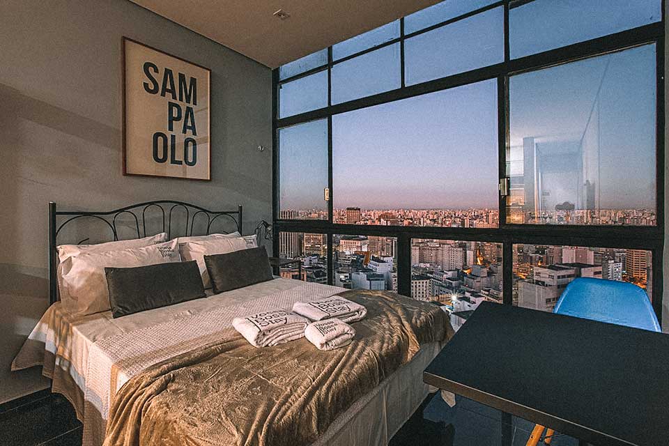 Cama de casal ao lado de grandes janelas com vista para o skyline de SP