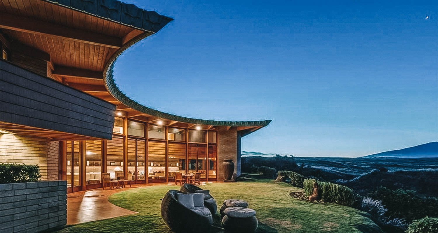 Área externa da casa com gramado e vista para as montanhas. Do lado esquerdo, parte da casa em madeira, iluminada e com grandes janelas de vidro