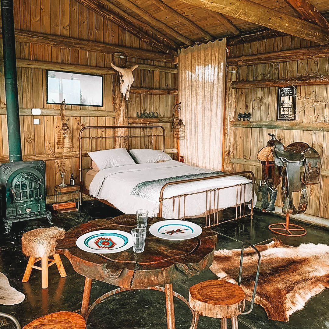 Quarto rústico com cama ao centro, banquinhos, mesa e parede de madeira
