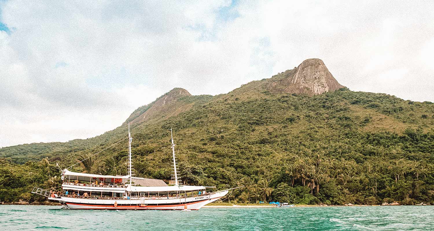 Barco de tamanho médio com Morro do Pico ao fundo, navegando pelas águas do Saco do Mamanguá, em Paraty-Mirim