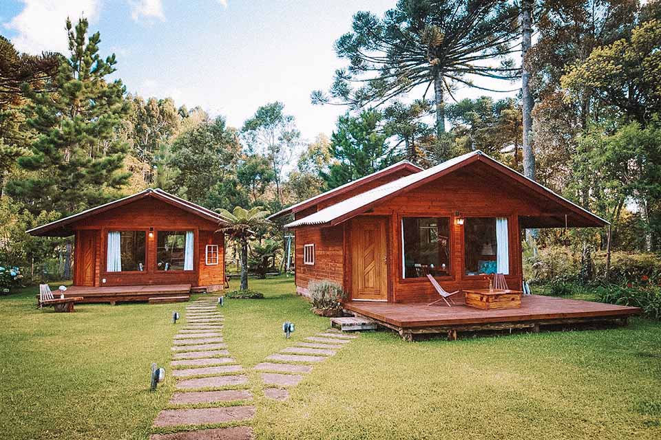 Cabanas de madeira em um espaço verde rodeado de araucárias, com caminho de pedra entre elas