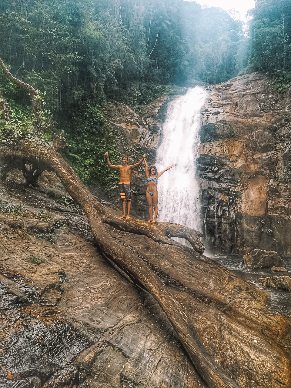 Um homem e uma mulher de braços abertos em frente à Cachoeira Boiçuncanga, perto de Maresias, em São Paulo