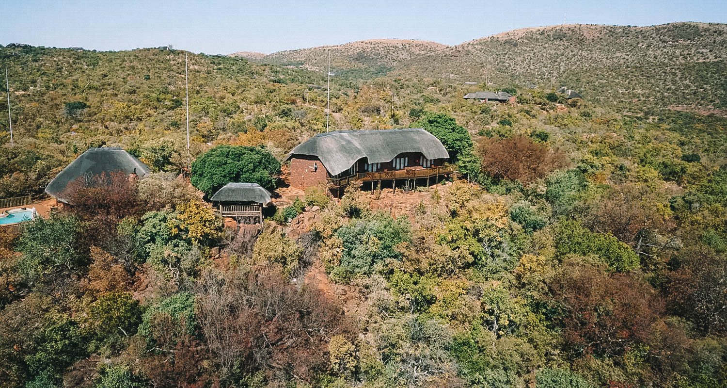 Imagem aérea que mostra a casa em madeira com deck envolto pot muitas árvores e montanhas