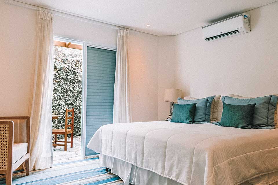 Quarto do hotel Maui Maresias: paredes brancas, cama com lençol branco e almofadas verdes e porta aberta com vista para jardim vertical