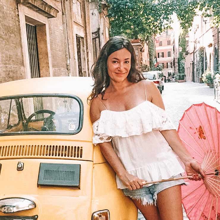 Mulher morena ao lado de um fusca amarelo, com uma blusa branca e short jeans, segurando uma sombrinha rosa. Juju na Trip Blog. 