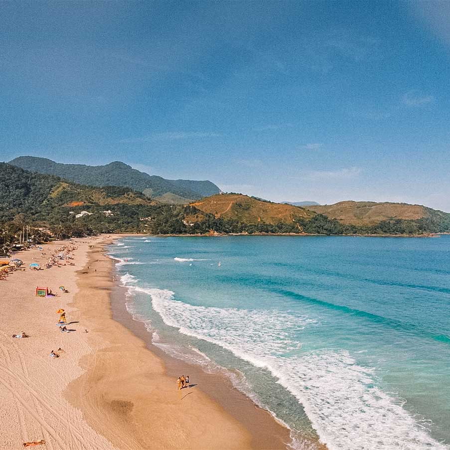 Vista do litoral de Maresias, com o mar azulado, pessoas na praia e a Serra do Mar ao fundo