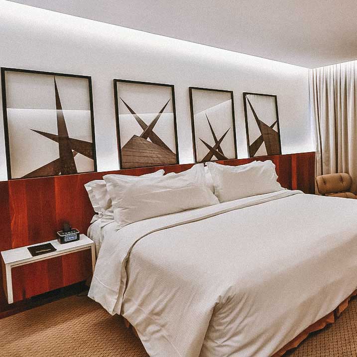 Quarto de hotel, com cama de casal e lençóis brancos, quatro quadros com paisagens de Goiânia e piso de madeira
