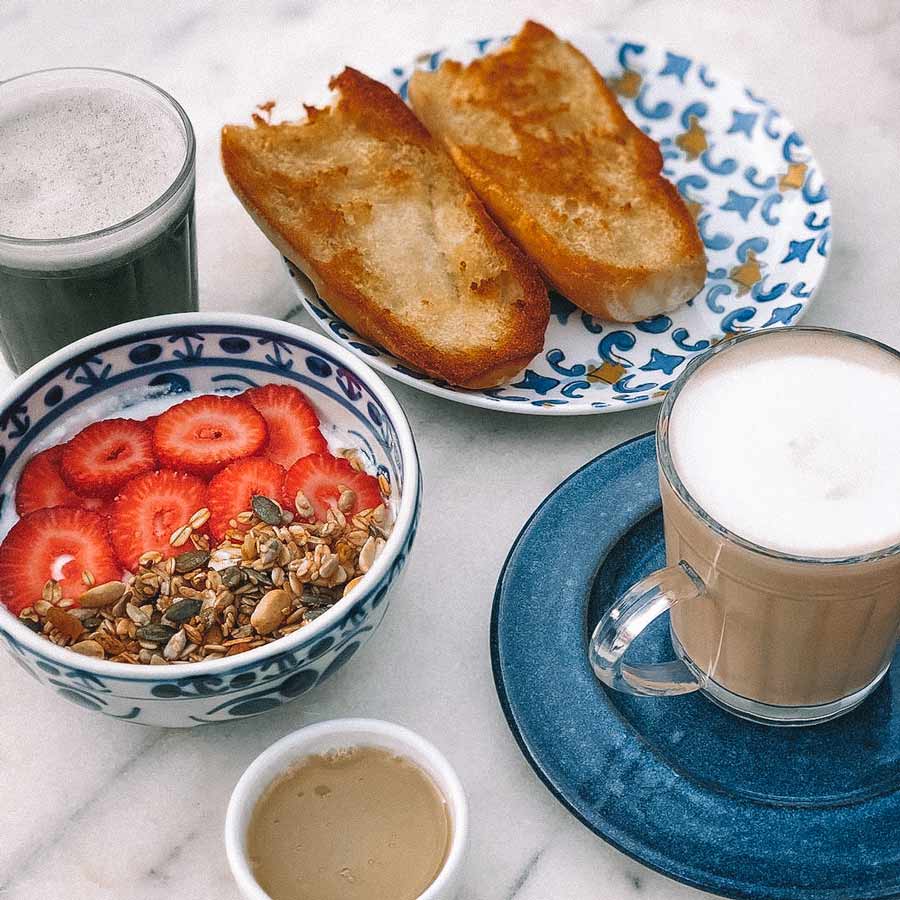 Bowl com morango e iogurte, pão na chapa com manteiga e diferentes cafés expostos numa mesa de mármore