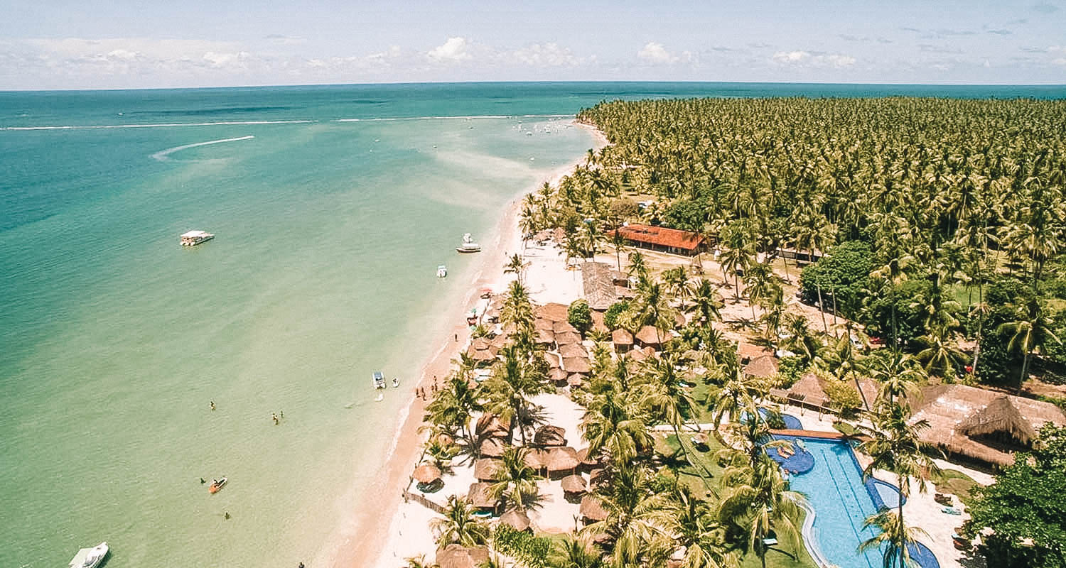 Vista aérea da divisa entre o mar em tom azulado, a areia e a pousada coberta por uma vegetação muito verde