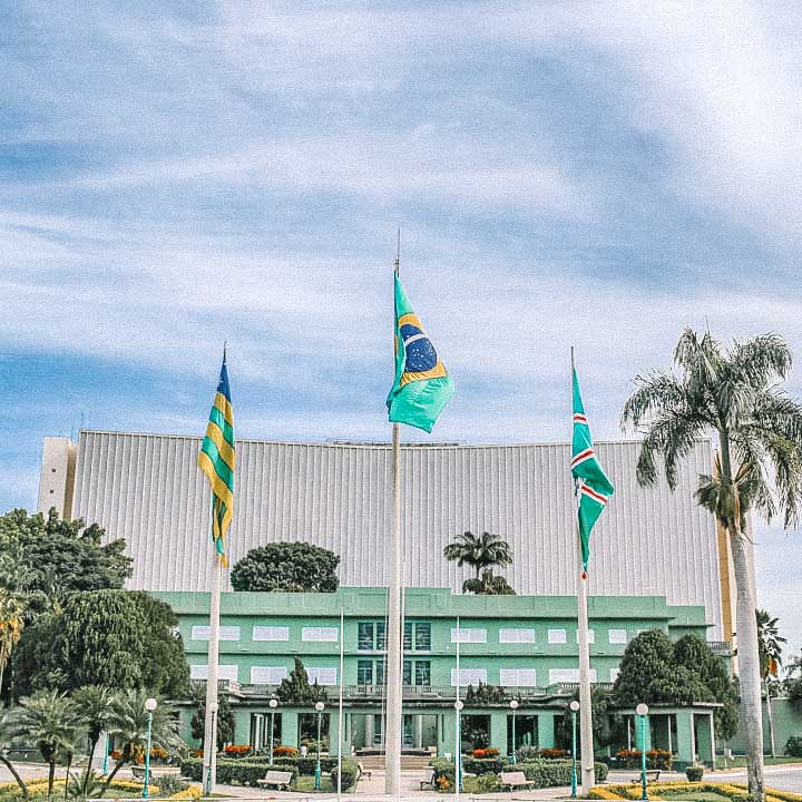 Três bandeiras hasteadas em frente a um edifício baixo, com árvores ao redor