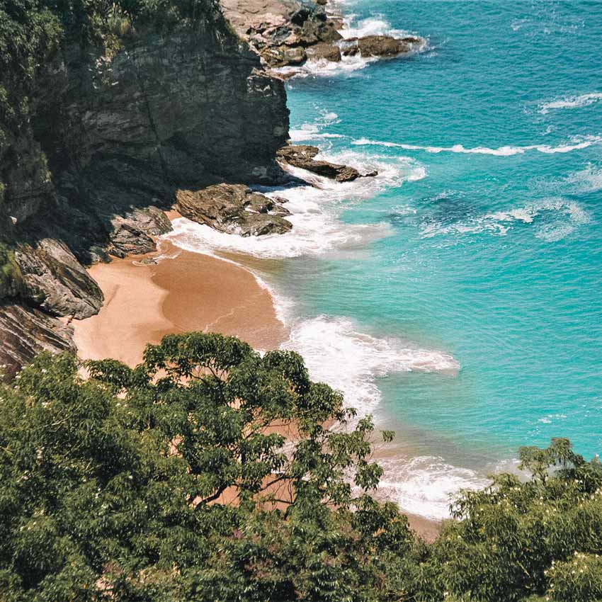 Praia cercada de pedras e vegetação com mar em tons de azul, no litoral Norte de São Paulo. Praia Brava - Maresias (SP).