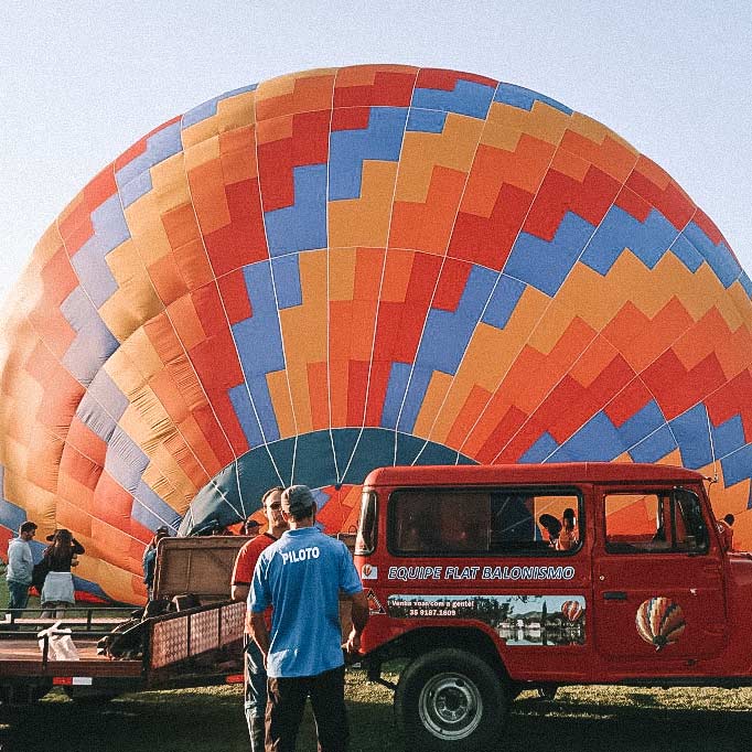 Em primeiro plano, um jipe vermelho e pessoas de costas olhando o balão colorido, em segundo plano, sendo montado. 