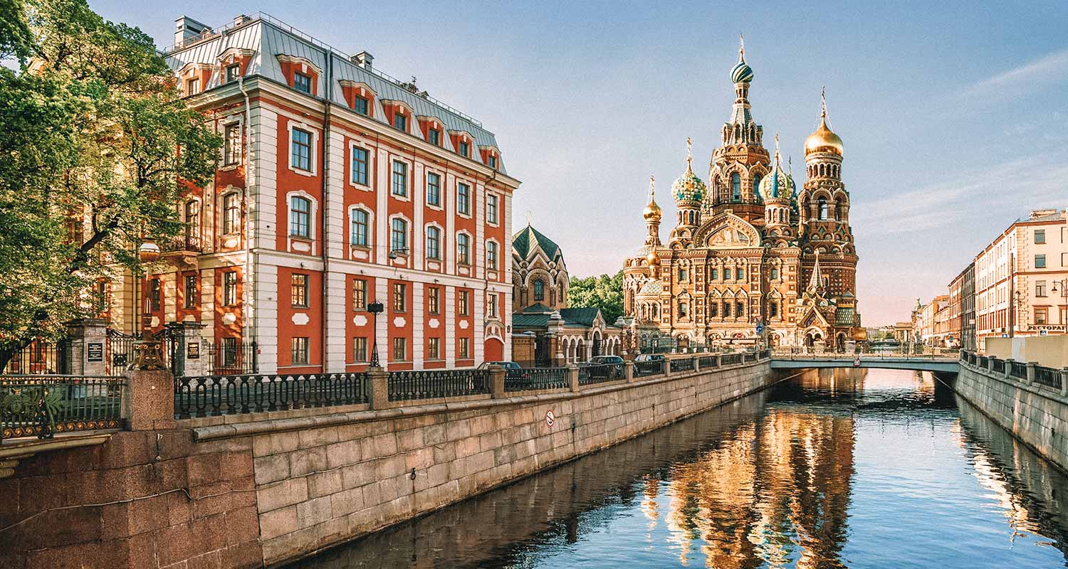 O canal de São Petersburgo, com um prédio vermelho ao lado, e uma catedral ao fundo. São Petersburgo é uma das maiores cidades da Europa.