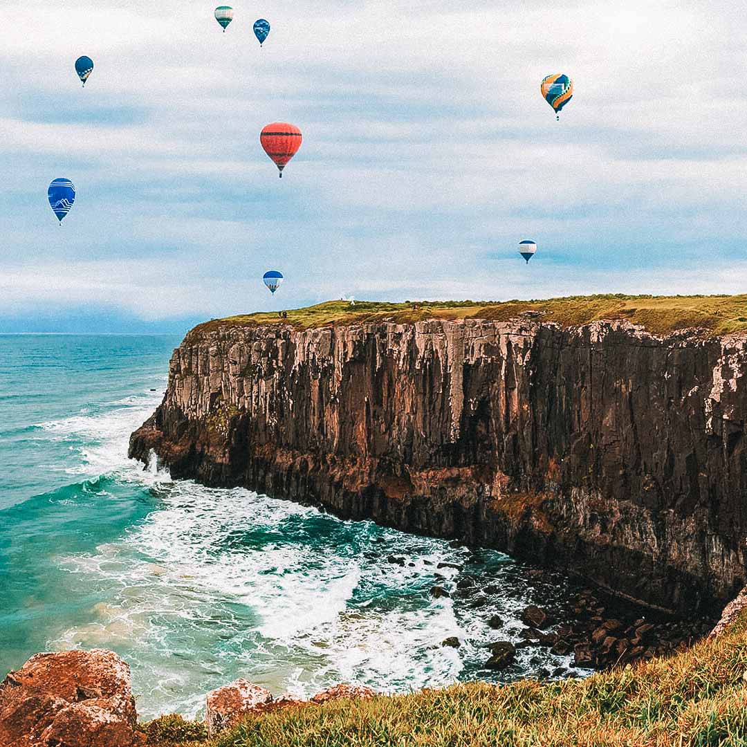 Vários balões no horizonte distante. Em primeiro, uma falésia e o mar azul-esverdeado batendo nas pedras. 