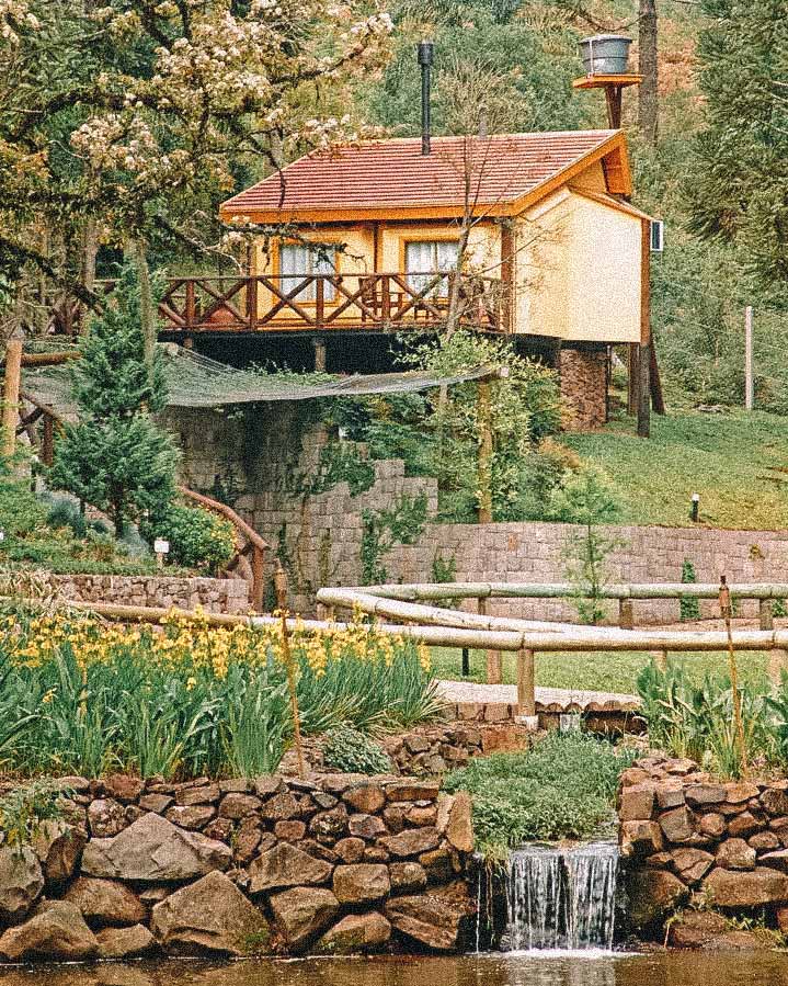 Bangalô suspenso amarelo, rodeado de plantas em segundo plano. Em primeira plano, um córrego com pequena cascata. Villa Flor Ecoresort em Nova Petrópolis