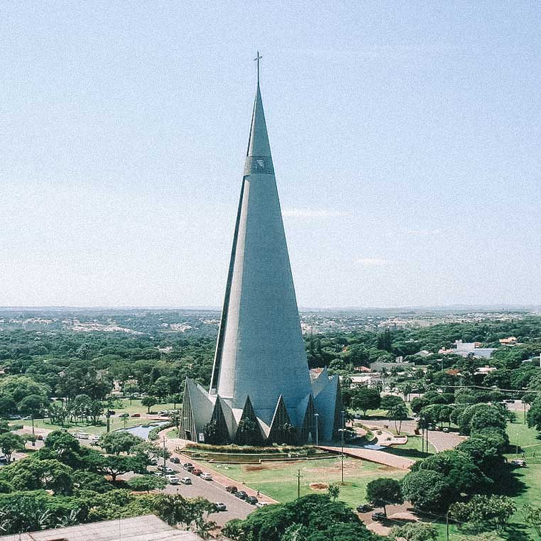 Igreja em formato cônico, em terreno plano, rodeada por várias árvores