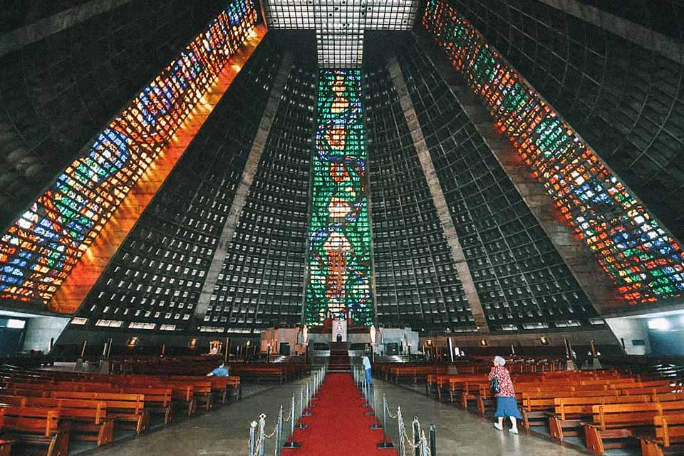 Catedral em formato cônico, com vitrais coloridos, tapete vermelho levando ao altar e bancos de madeira