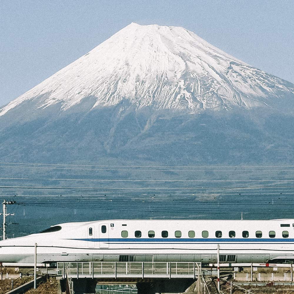 Trem branco passando em ferrovia, com uma montanha de cume enevoado atrás. Trens mais rápidos do mundo.