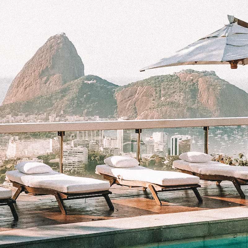 Espreguiçadeiras de madeira com almofadas brancas em rooftop com vista para o Pão de Açúcar