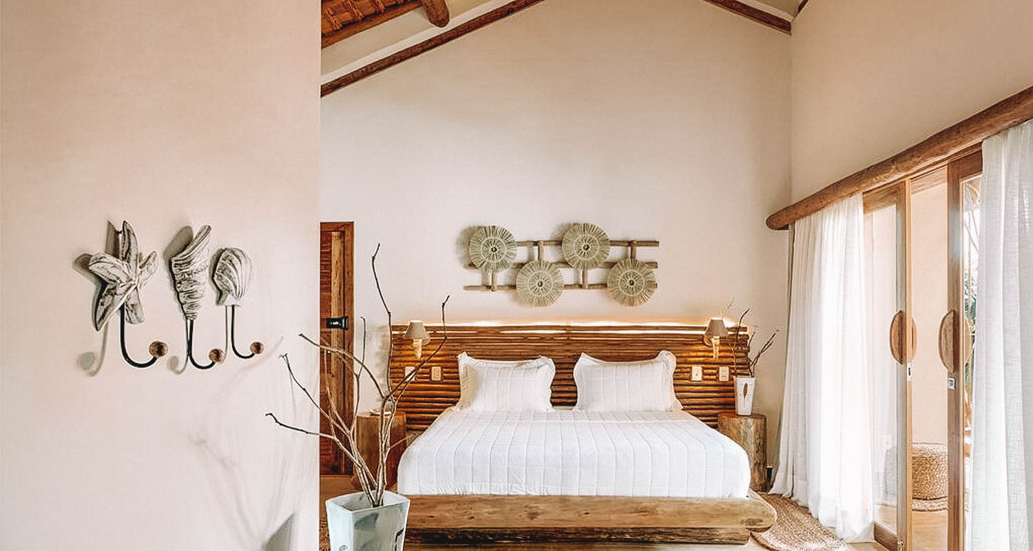 Quarto amplo com paredes brancas, decoração em palha e madeira e cama de casal ao meio