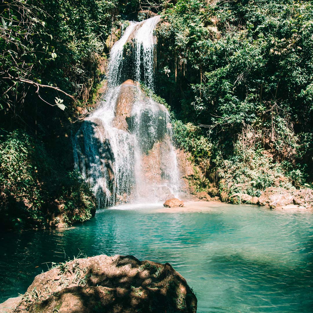 Poço com água de coloração azul, rochas ao redor e uma grande cascata ao fundo